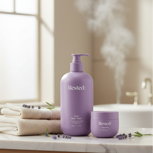Purple 'Rested' body wash and cream on a bathroom counter with towels and lavender flowers.